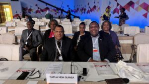 Group photo of 6 individuals at an international conference, seated behind a country name sign saying Papua New Guinea