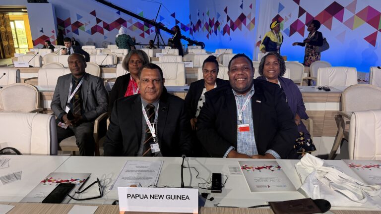 Group photo of 6 individuals at an international conference, seated behind a country name sign saying Papua New Guinea